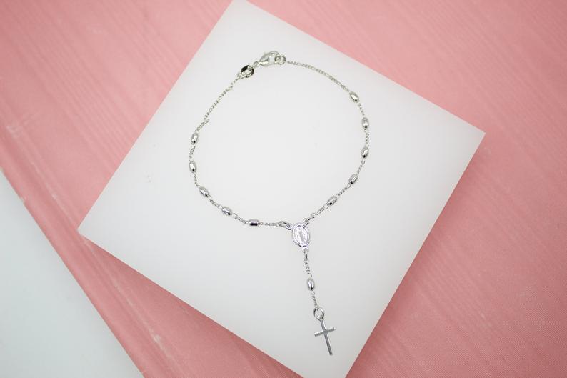 A delicate silver rosary bracelet featuring small beads and a hanging cross rests on a white display against a pink background.