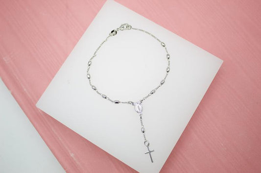 A delicate silver rosary bracelet featuring small beads and a hanging cross rests on a white display against a pink background.