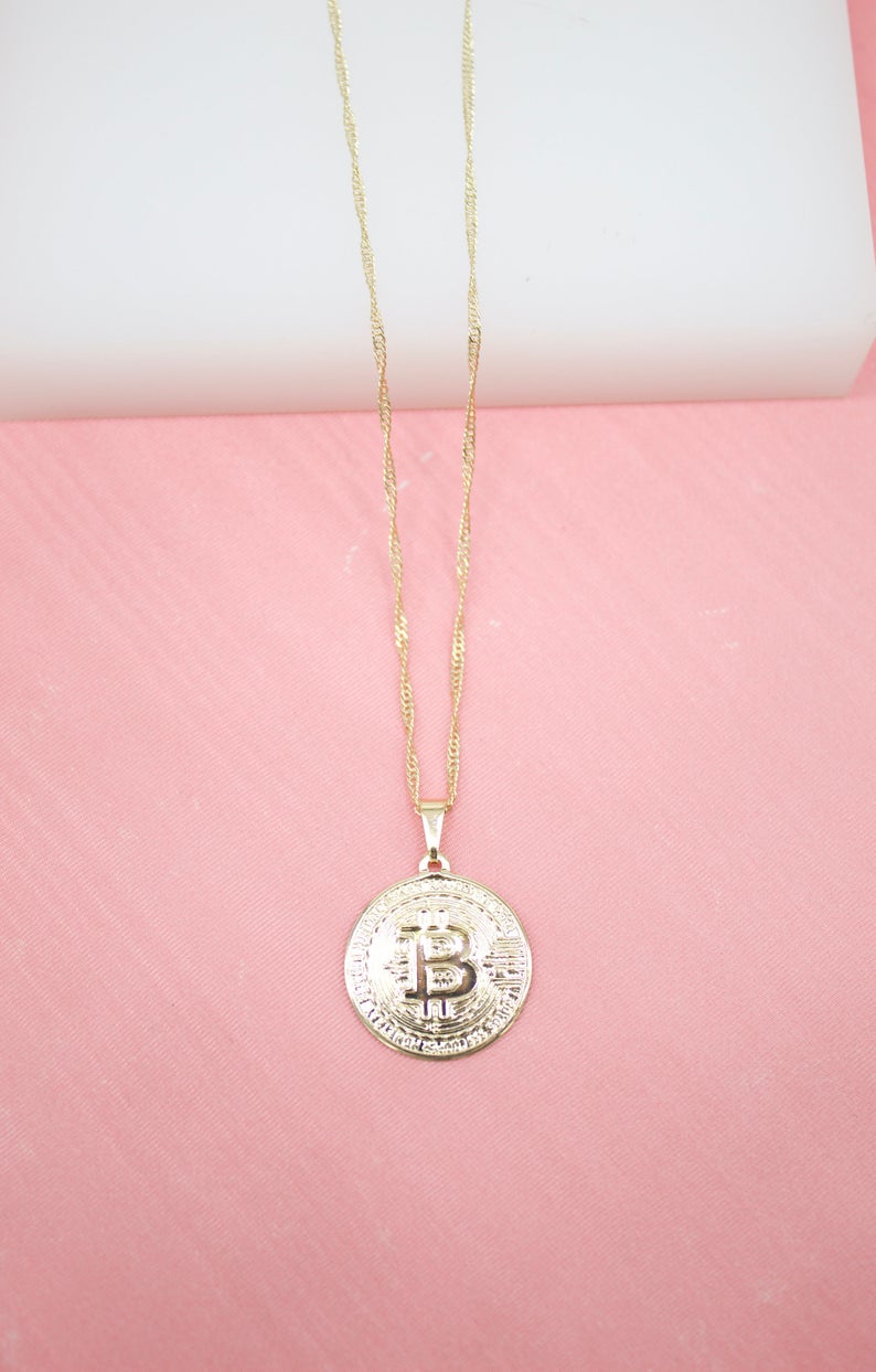 A delicate gold chain holds a textured Bitcoin coin pendant against a soft pink background.