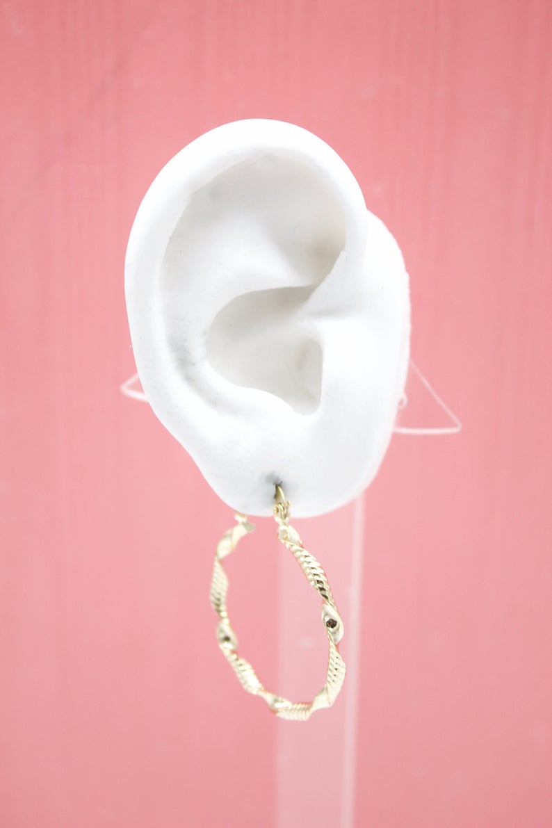 A white mannequin ear displays a stylish textured gold hoop earring against a soft pink background.