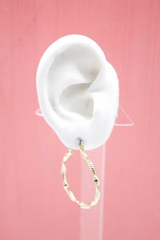 A white mannequin ear displays a textured gold hoop earring against a pink background.