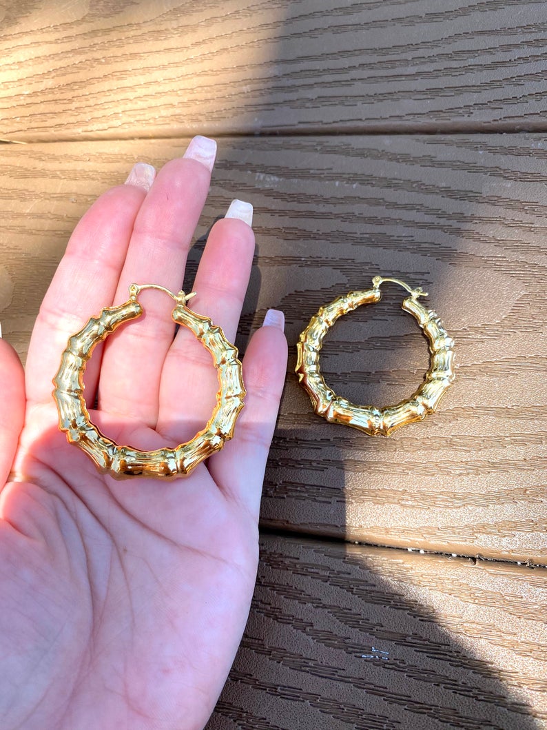 A person's hand holds a pair of shiny gold bamboo hoop earrings against a dark wooden surface.