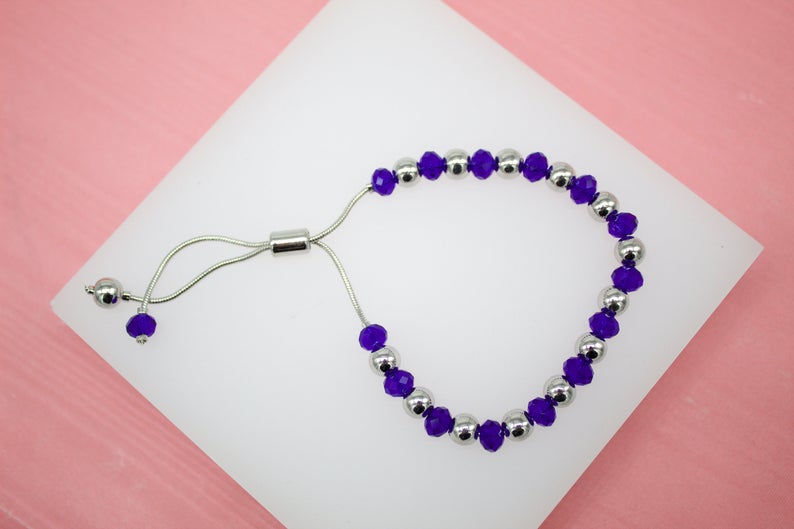 A delicate silver slider bracelet features alternating purple crystal beads and polished silver spheres, resting on a white display against a pink background.