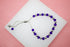 A delicate silver slider bracelet features alternating purple crystal beads and polished silver spheres, resting on a white display against a pink background.