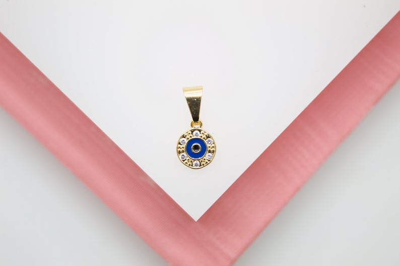 A gold evil eye pendant featuring blue enamel and sparkling zirconia stones rests centrally on a pristine white display surface framed by soft pink acrylic edges.