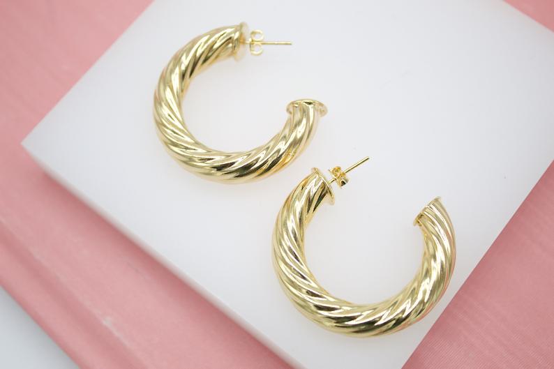 A pair of glossy gold twisted hoop earrings rests on a white display stand.