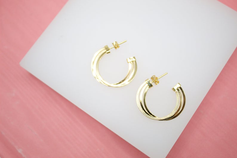 A pair of gold multi-band hoop earrings rests on a white display against a pink background.