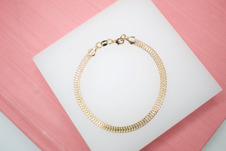 A gold herringbone chain bracelet rests gracefully on a white display block against a soft pink background.