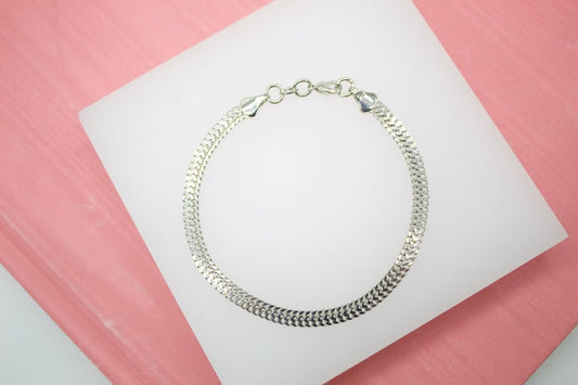 A sleek silver herringbone chain bracelet rests on a white display pad against a pink background.