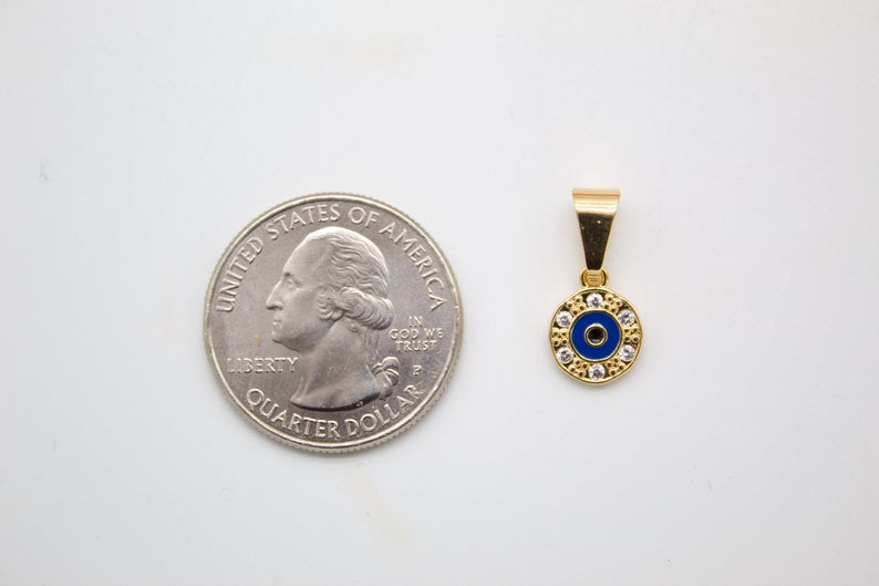 A tiny gold evil eye pendant with blue enamel and cubic zirconia stones sits next to a US quarter for size comparison on a white surface.