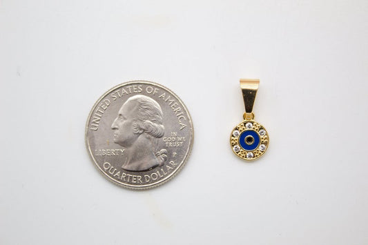 A tiny gold evil eye pendant with blue enamel and cubic zirconia stones sits next to a US quarter for size comparison on a white surface.