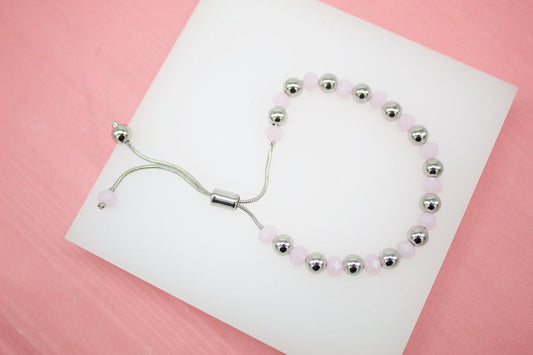 A silver adjustable slider bracelet features alternating shiny metal beads and soft pink crystal stones resting on a white display against a pink background.