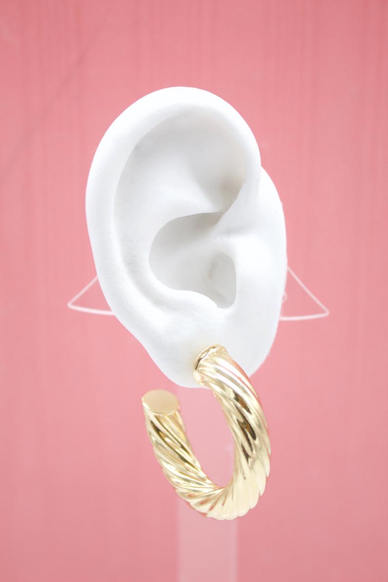 A white mannequin ear displays a chunky, twisted gold hoop earring against a soft pink background.
