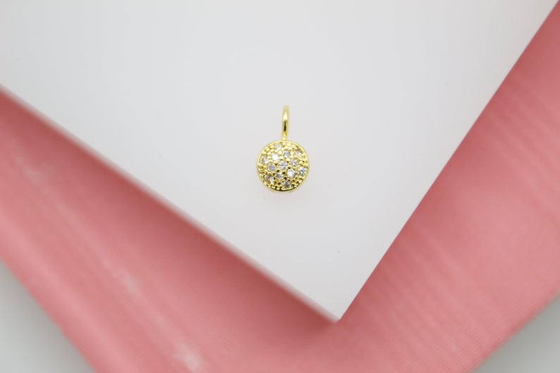 A sparkling gold pavé charm rests on a white display block against a soft pink background.
