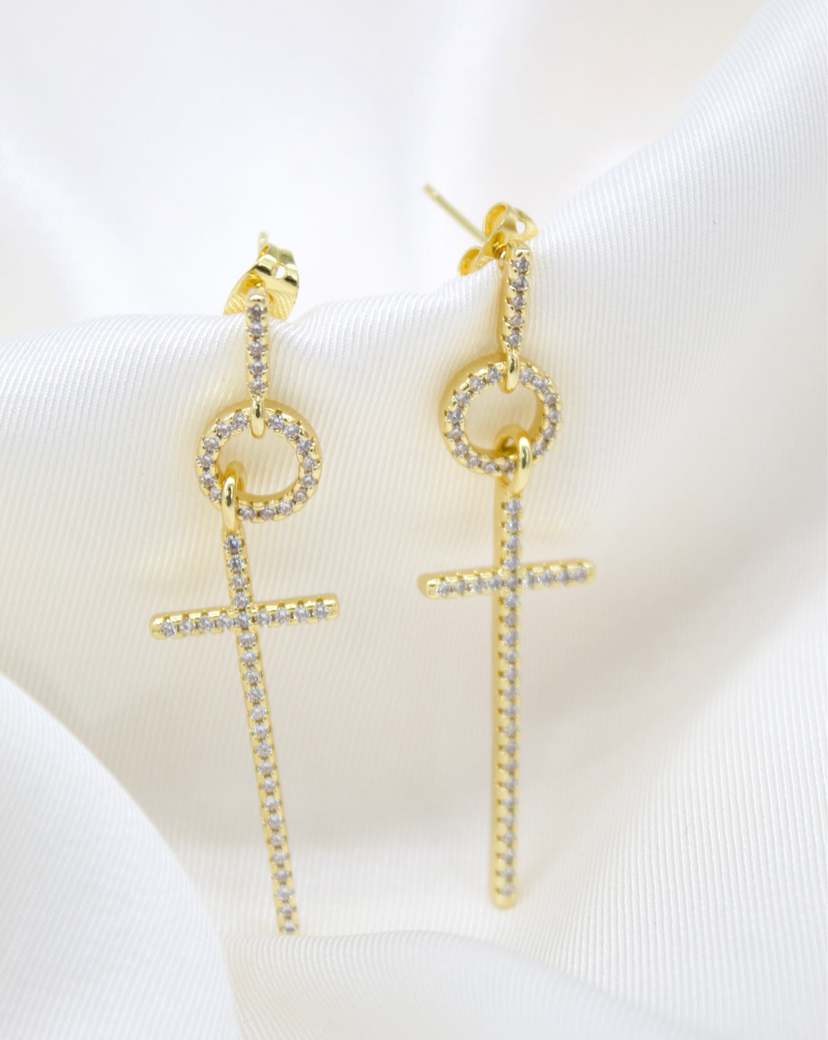 A pair of sparkling gold cross drop earrings featuring paved cubic zirconia stones rests elegantly on a soft white silk fabric.