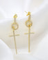 A pair of sparkling gold cross drop earrings featuring paved cubic zirconia stones rests elegantly on a soft white silk fabric.