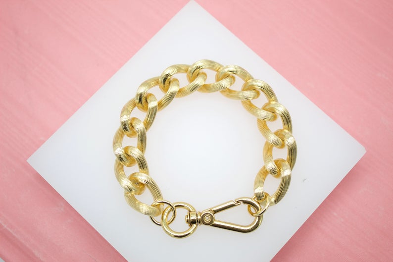 A textured gold chain bracelet rests on a white square display card against a pink background.