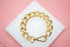 A textured gold chain bracelet rests on a white square display card against a pink background.