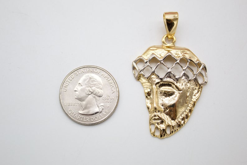 A two-tone gold Jesus head pendant rests next to a US quarter coin to show its scale.
