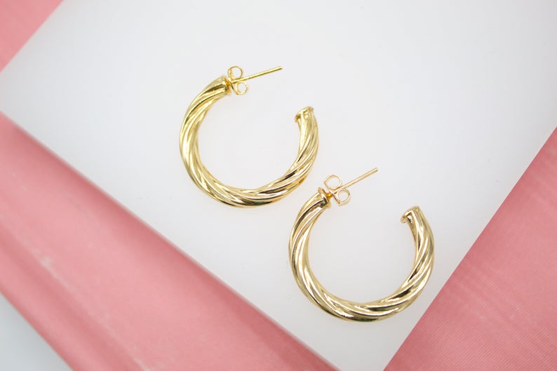 A pair of polished gold twist hoop earrings rests elegantly on a white surface against a pink backdrop.