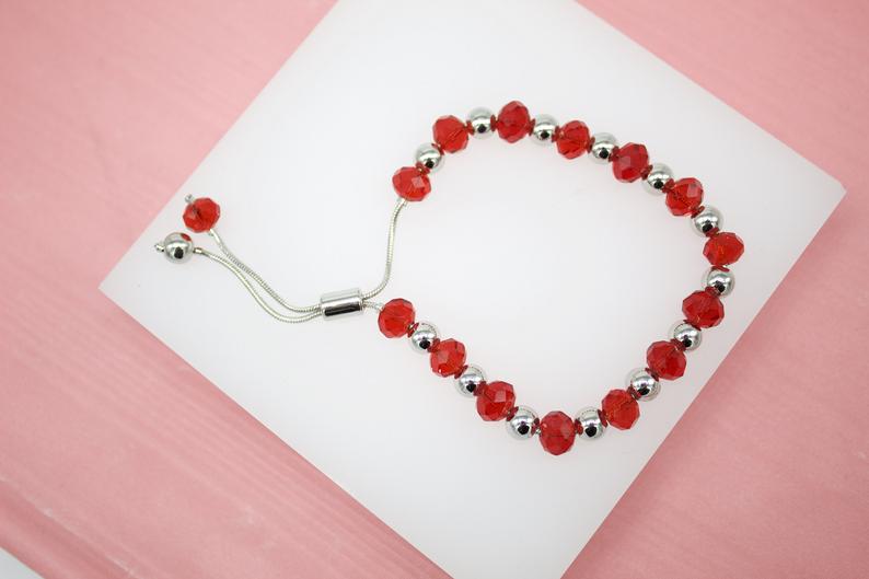 A delicate silver adjustable bracelet features alternating red crystal beads and metallic balls displayed on a white surface against a pink background.