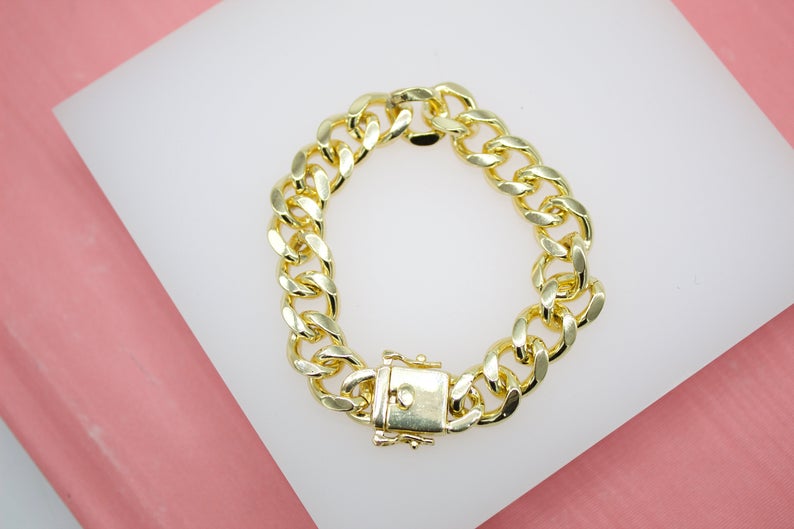 A thick gold-plated curb chain bracelet rests on a white display surface against a pink background.