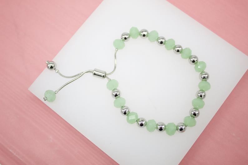 An adjustable silver slider bracelet features alternating light green crystal beads and shiny metallic silver balls.