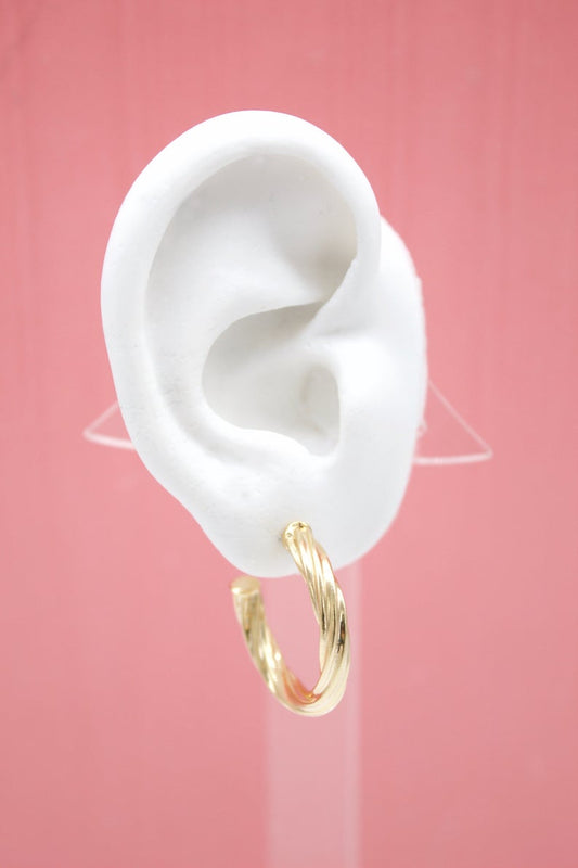 A white mannequin ear displays a textured, twisted gold hoop earring against a solid pink background.