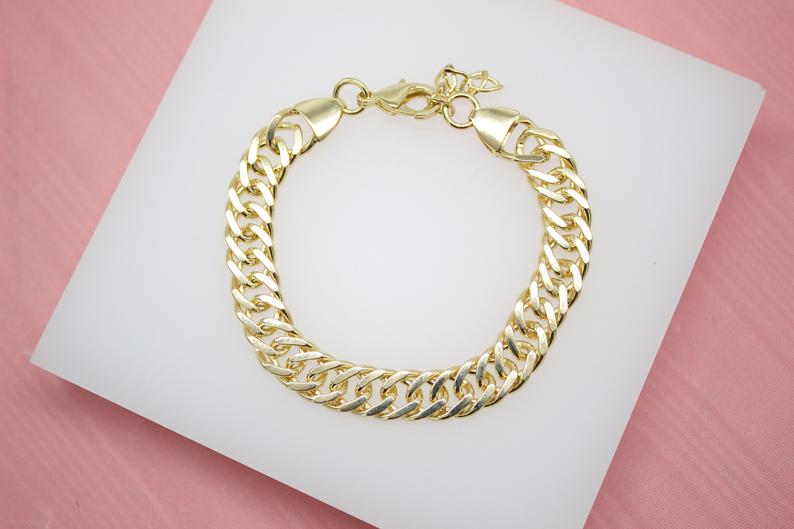 A shiny gold chain bracelet rests on a white display pad against a pink background.