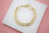 A shiny gold chain bracelet rests on a white display pad against a pink background.