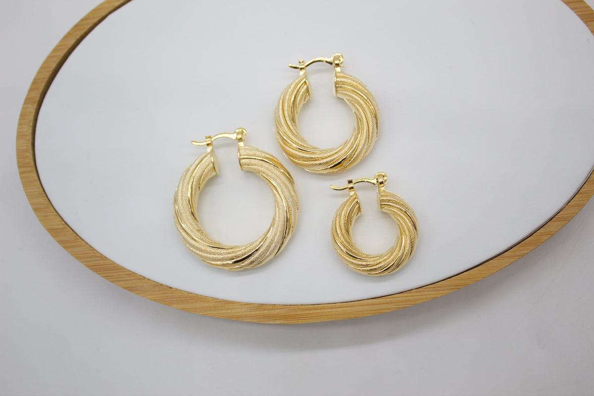 Three textured gold hoop earrings in descending sizes rest neatly on a white display tray with a wooden rim.