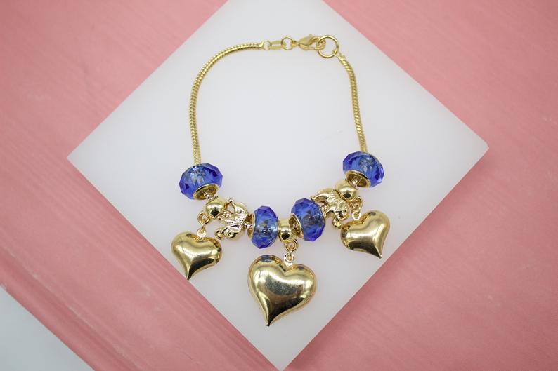 A gold charm bracelet featuring sparkling blue beads and dangling heart charms rests on a white display against a pink background.