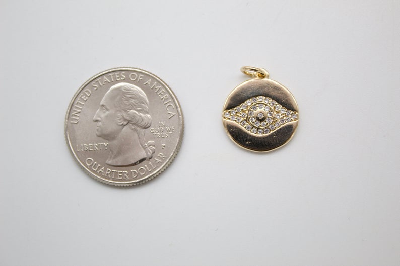 A small gold evil eye pendant with pavé diamonds rests next to a US quarter for scale.