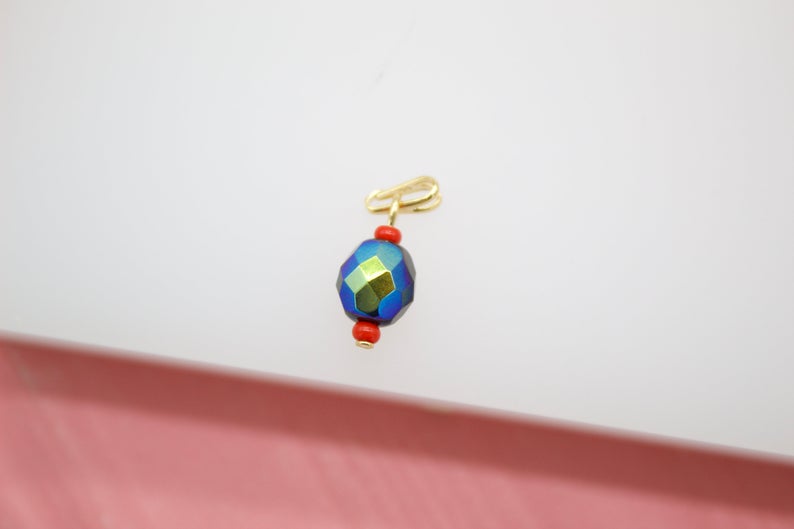 A gold-plated charm features a faceted iridescent blue bead flanked by two small red beads against a light background.