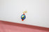 A gold-plated charm features a faceted iridescent blue bead flanked by two small red beads against a light background.