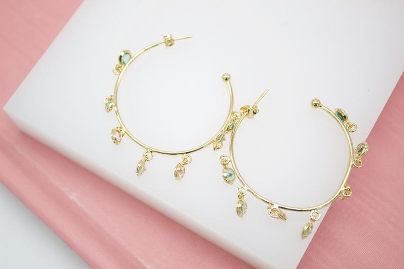 A pair of gold hoop earrings features colorful dangling gemstone charms against a white and pink background.