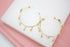 A pair of gold hoop earrings features colorful dangling gemstone charms against a white and pink background.