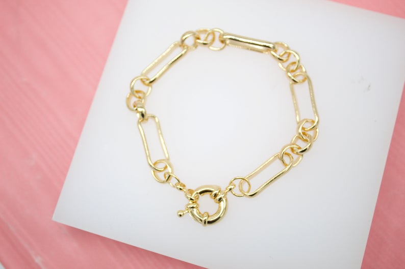 A shiny gold paperclip chain bracelet rests on a white display surface.