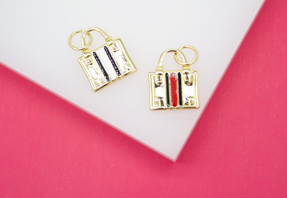 Two gold handbag charms with enamel stripes rest on a white surface against a bright pink background.