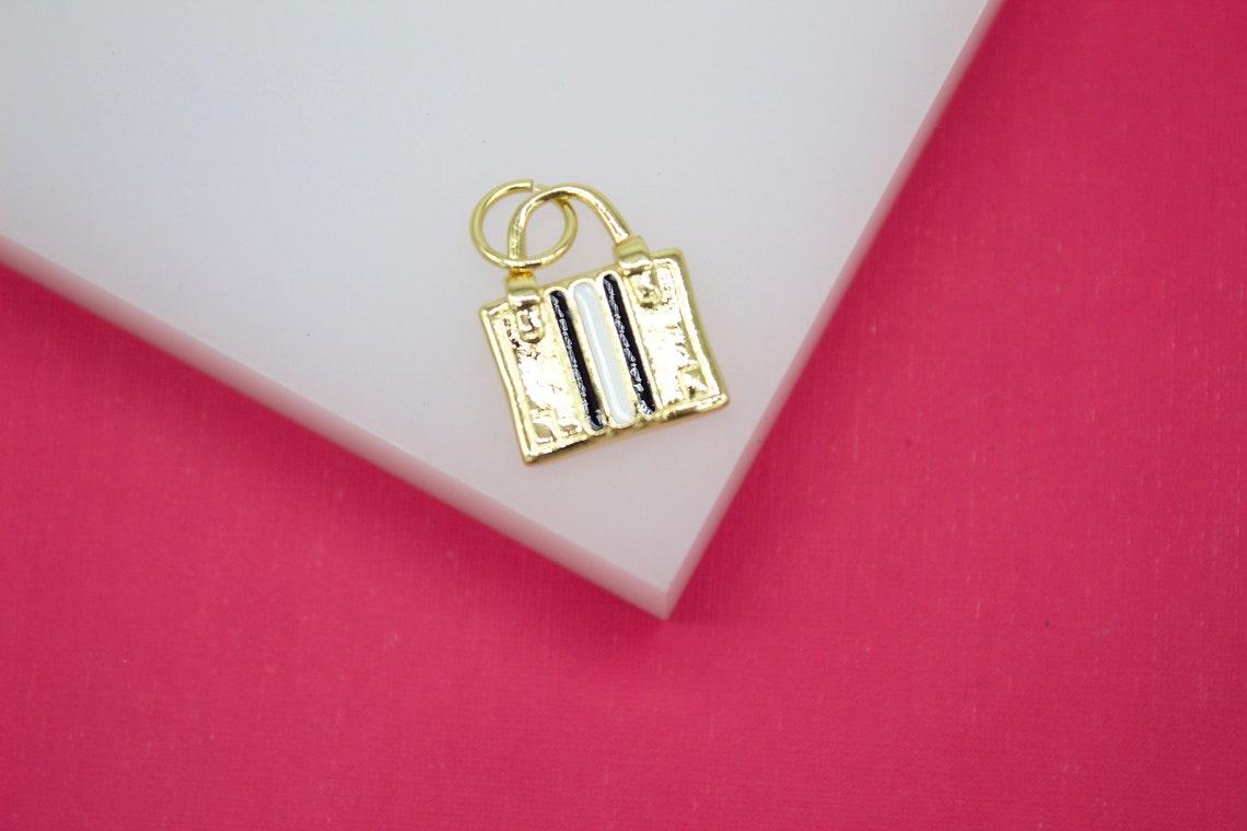 A gold handbag charm featuring black and white vertical stripes rests on a white display surface against a vibrant pink background.
