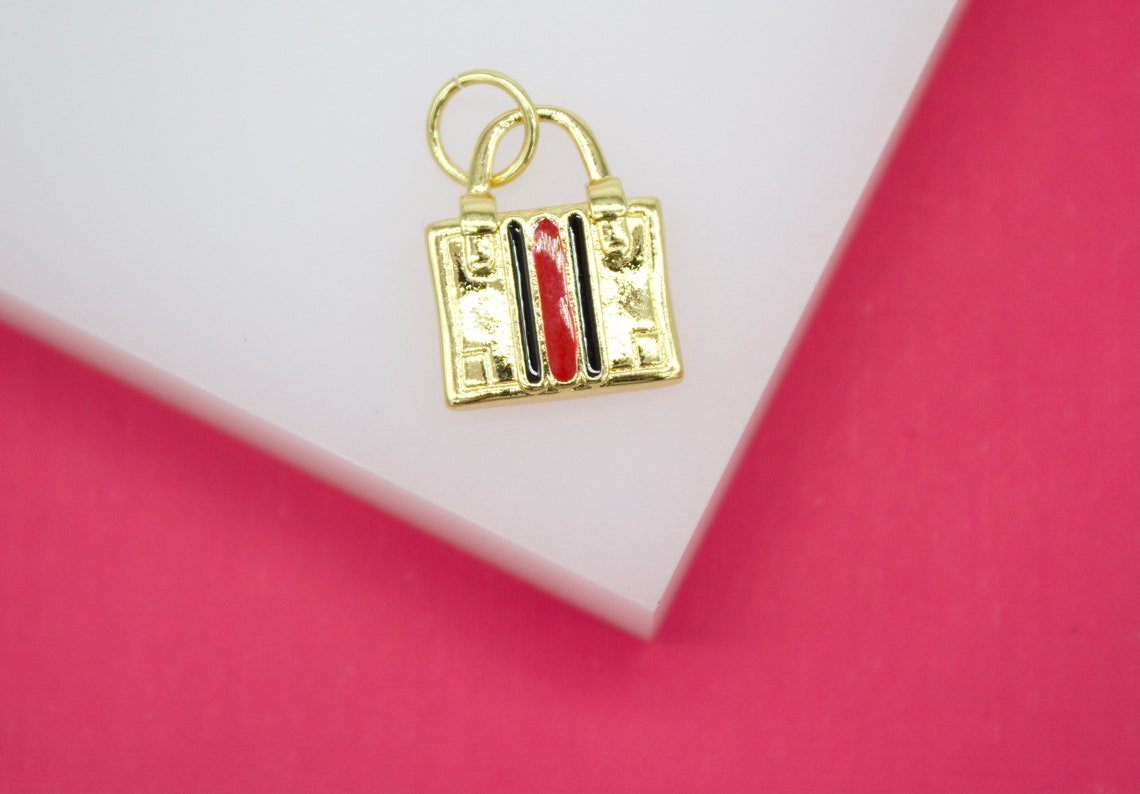 A gold handbag charm featuring red and black enamel stripes rests on a white display surface against a pink background.