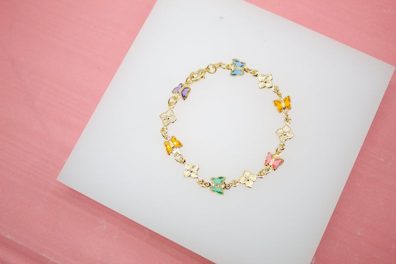 A gold children's bracelet features colorful enamel butterflies alternating with small flowers, displayed on a white surface against a pink background.
