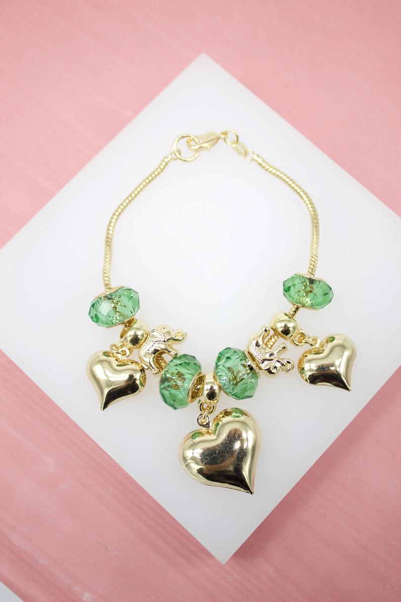 A gold charm bracelet features faceted green crystal beads, shiny gold heart charms, and a small elephant figurine, all displayed on a white surface.