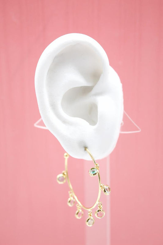 A white mannequin ear displays a gold charm hoop earring adorned with multicolored gemstone drops against a pink backdrop.