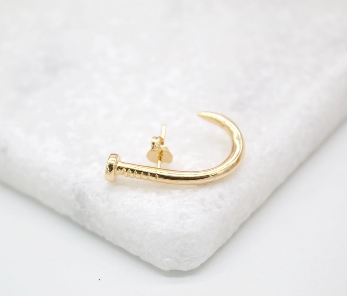 A gold nail-shaped stud earring rests on a textured white surface.