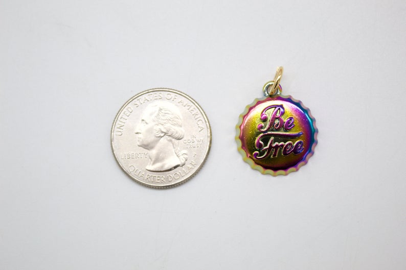 A rainbow-colored bottle cap charm with the words "Be Free" rests beside a US quarter for size comparison on a white surface.