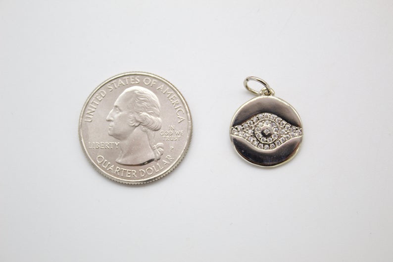 A silver evil eye pendant embellished with tiny sparkling cubic zirconia stones rests next to a US quarter coin for scale on a light gray surface.