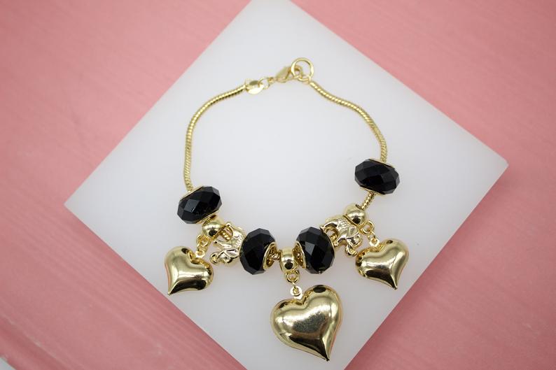 A gold charm bracelet featuring black crystal beads and dangling heart charms rests on a white display surface against a pink background.