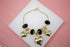 A gold charm bracelet featuring black crystal beads and dangling heart charms rests on a white display surface against a pink background.