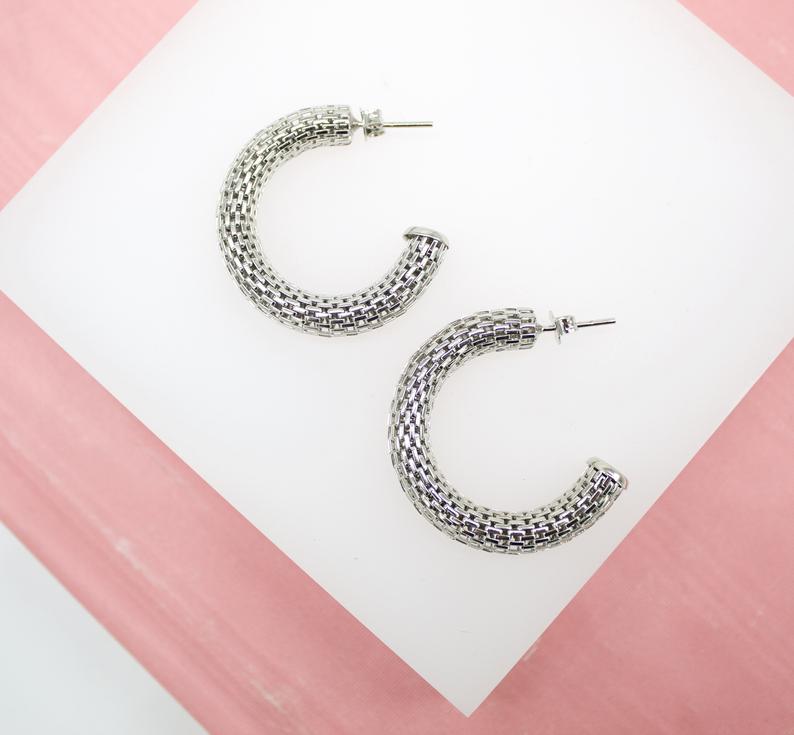 A pair of textured silver hoop earrings rests on a white display surface against a soft pink background.